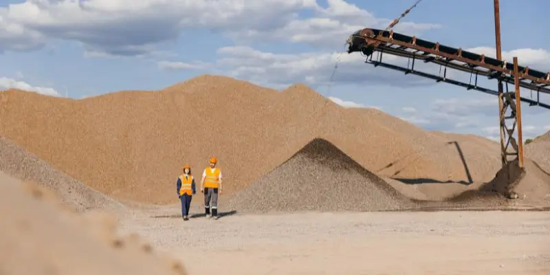 Two workers in construction gear stand before a large mound of crushed rock at a quarry site. Crusher troubleshooting.